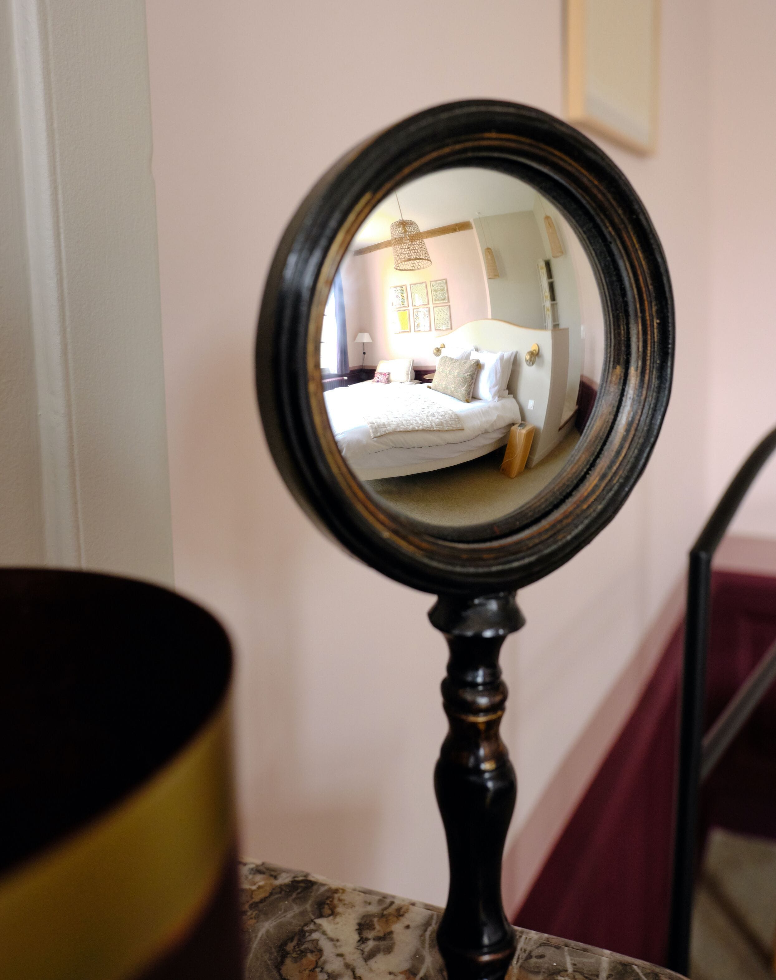 A round mirror on a stand, reflecting a bedroom with a bed, bedside table, and a wall with framed pictures.