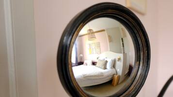 A round mirror on a stand, reflecting a bedroom with a bed, bedside table, and a wall with framed pictures.