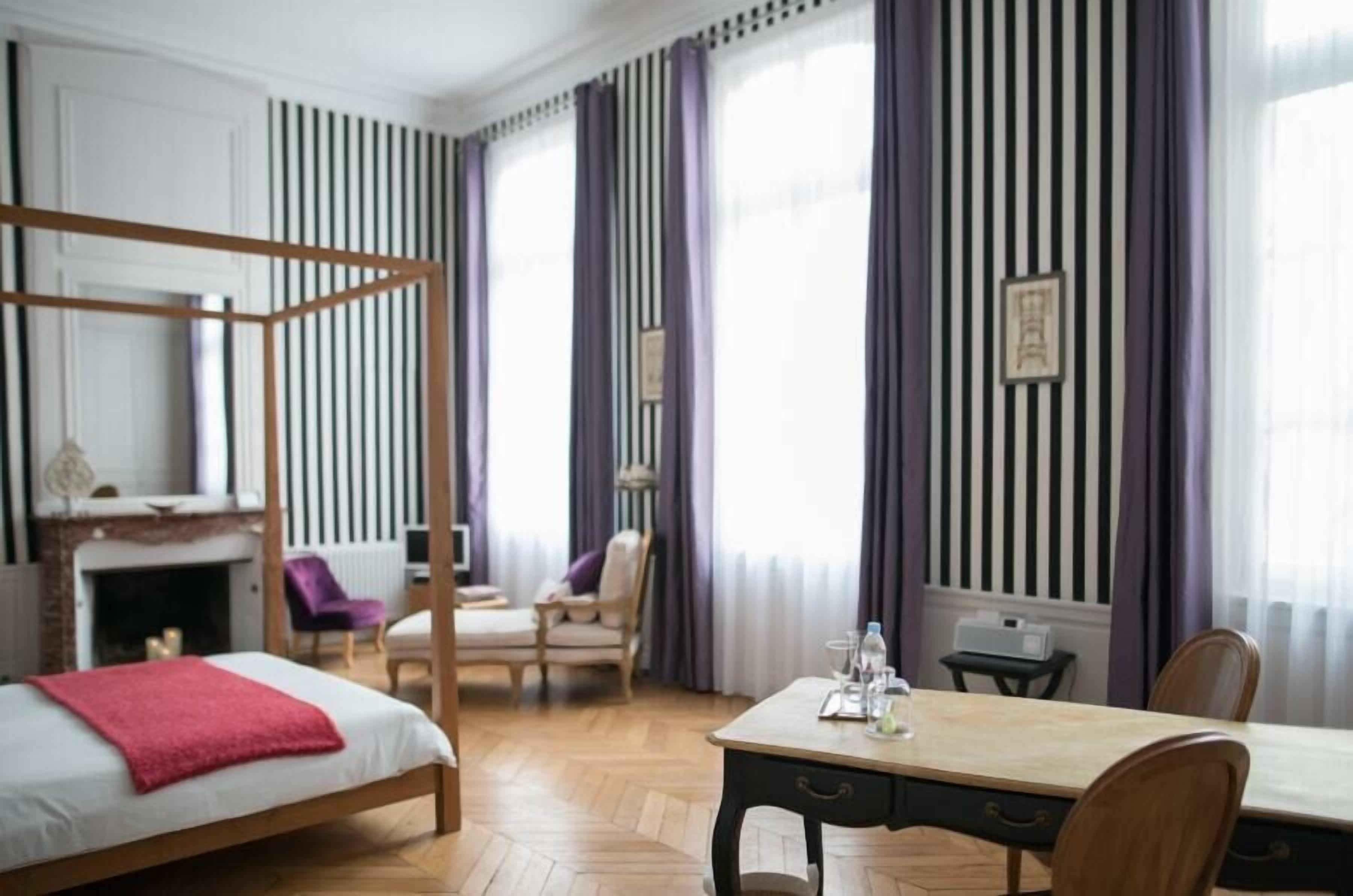 A hotel room with a four-poster bed, a wooden desk, a chair, a table with a bottle and glasses, and a fireplace.