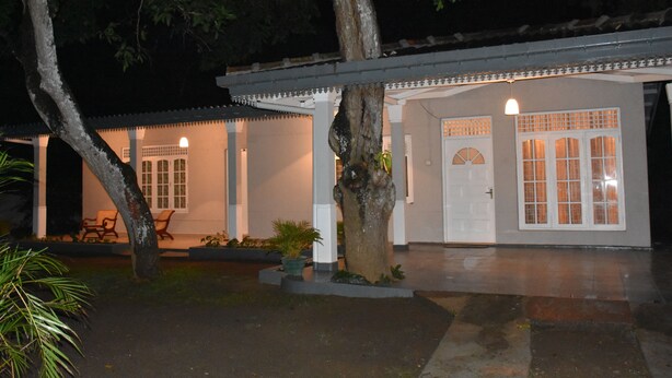 Un edificio tradicional con un porche cubierto, una puerta con paneles de vidrio y un pequeño jardín con sillas y plantas en macetas.