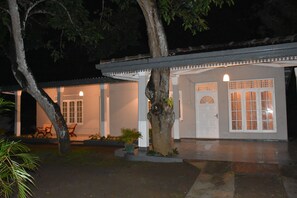 A traditional building with a covered porch, a door with glass panels, and a small garden area with chairs and potted plants.