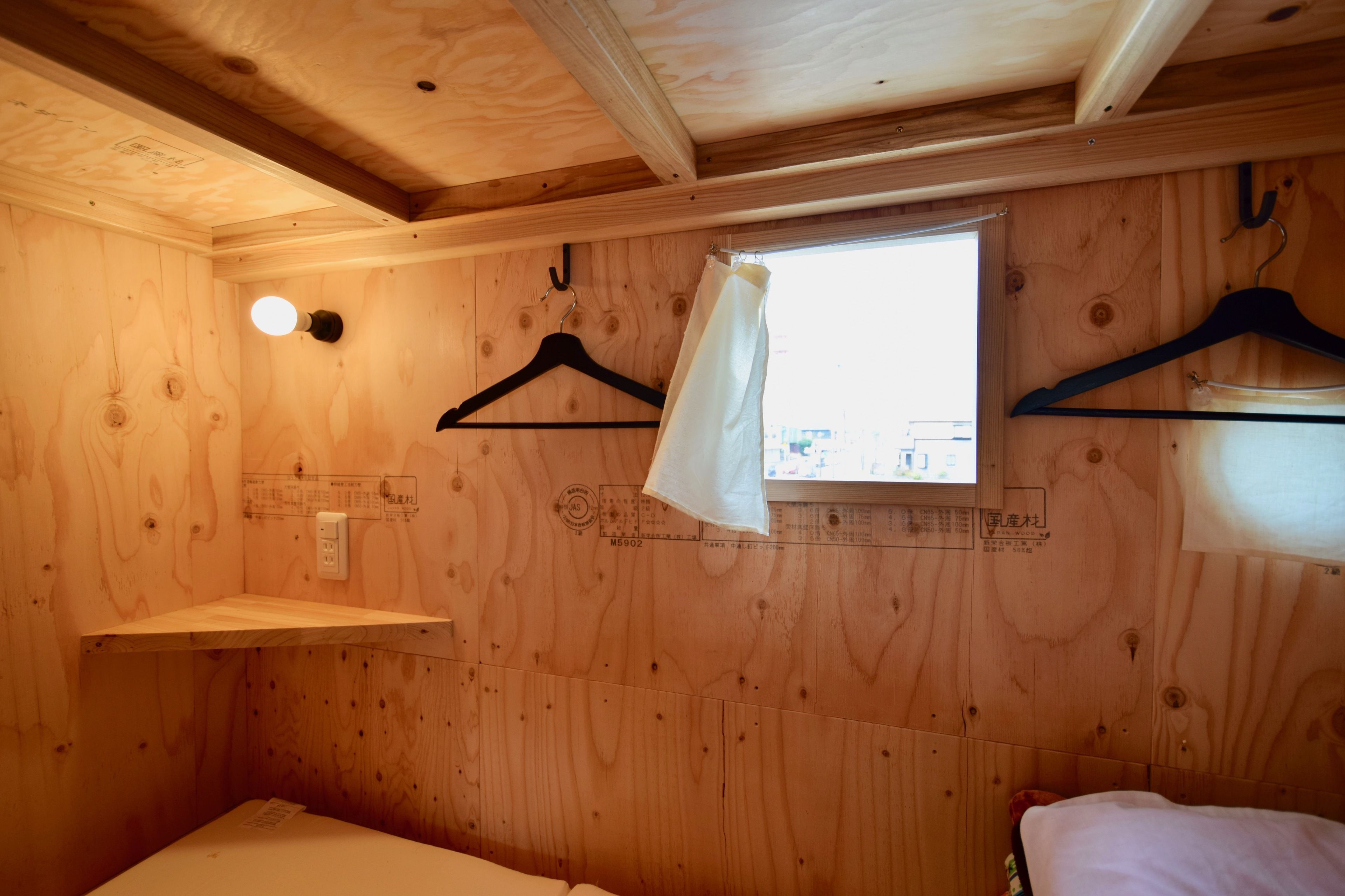 Una habitación de cabaña pequeña y de madera con una cama individual, una ventana y dos perchas negras en la pared.