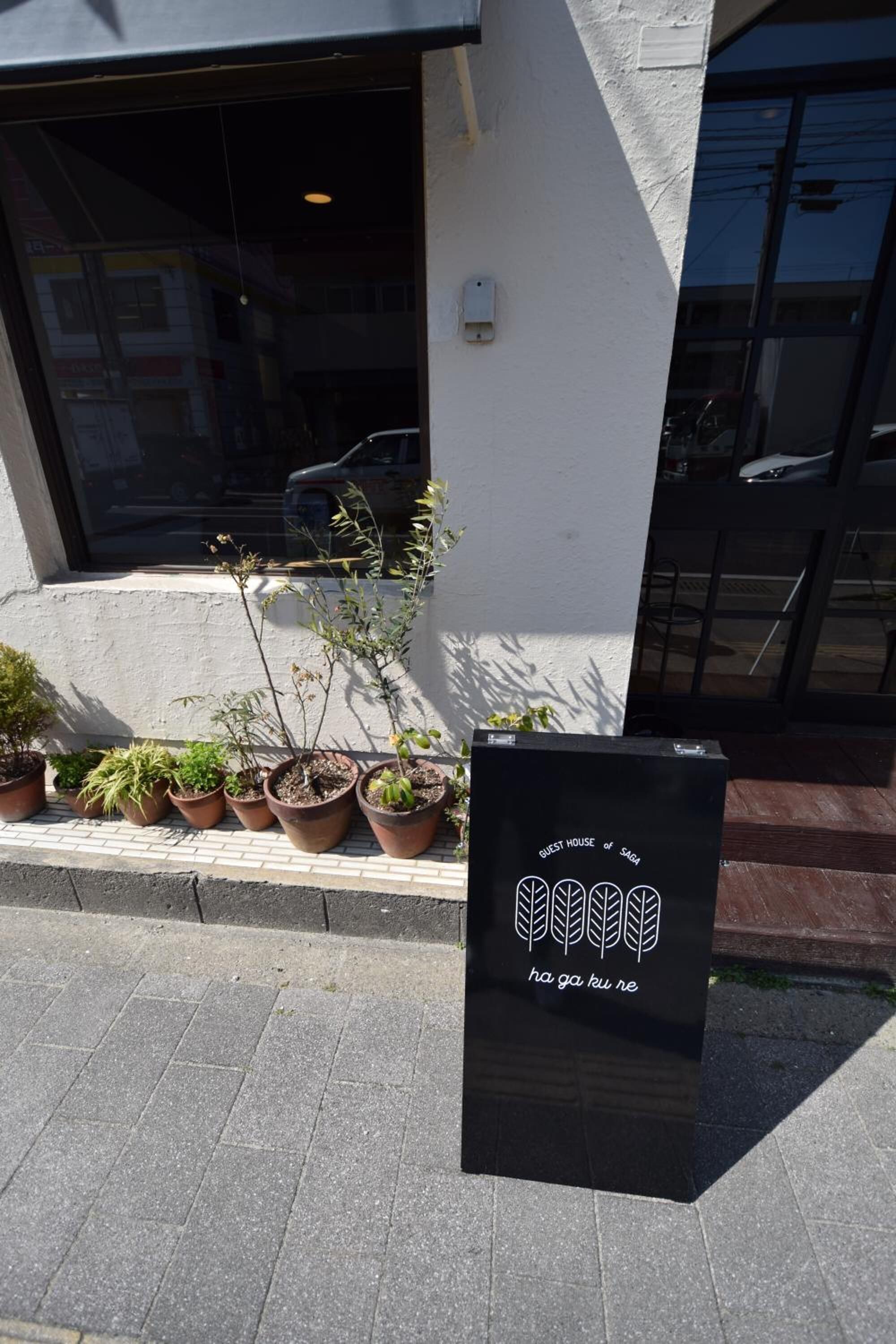 A black sign with white text and a logo, placed on a sidewalk next to a row of potted plants.