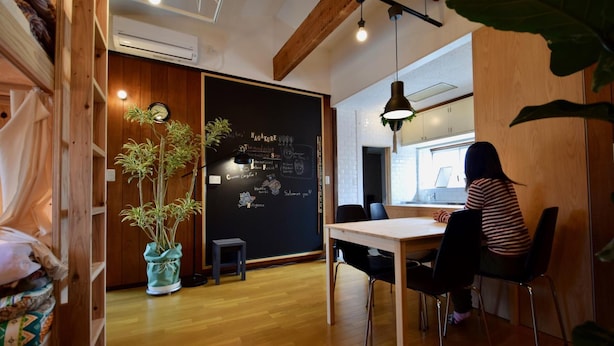 A cozy room with a wooden ceiling, a blackboard with writing, a potted plant, and a person sitting at a table. - SAGA International Guesthouse HAGAKURE - Hostel (Saga)