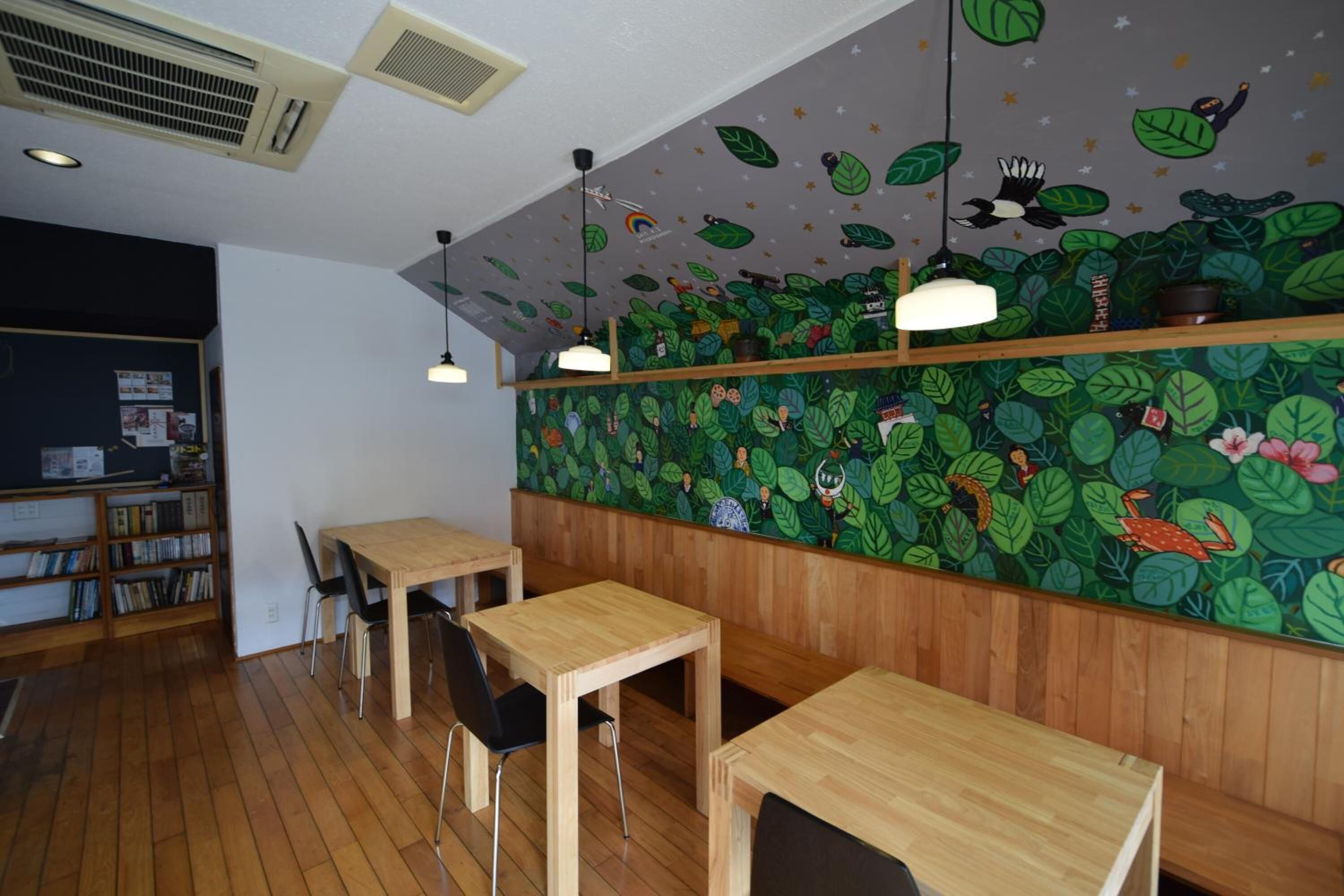 A restaurant with wooden tables and chairs, a green leafy wall mural, and a bookshelf with books.