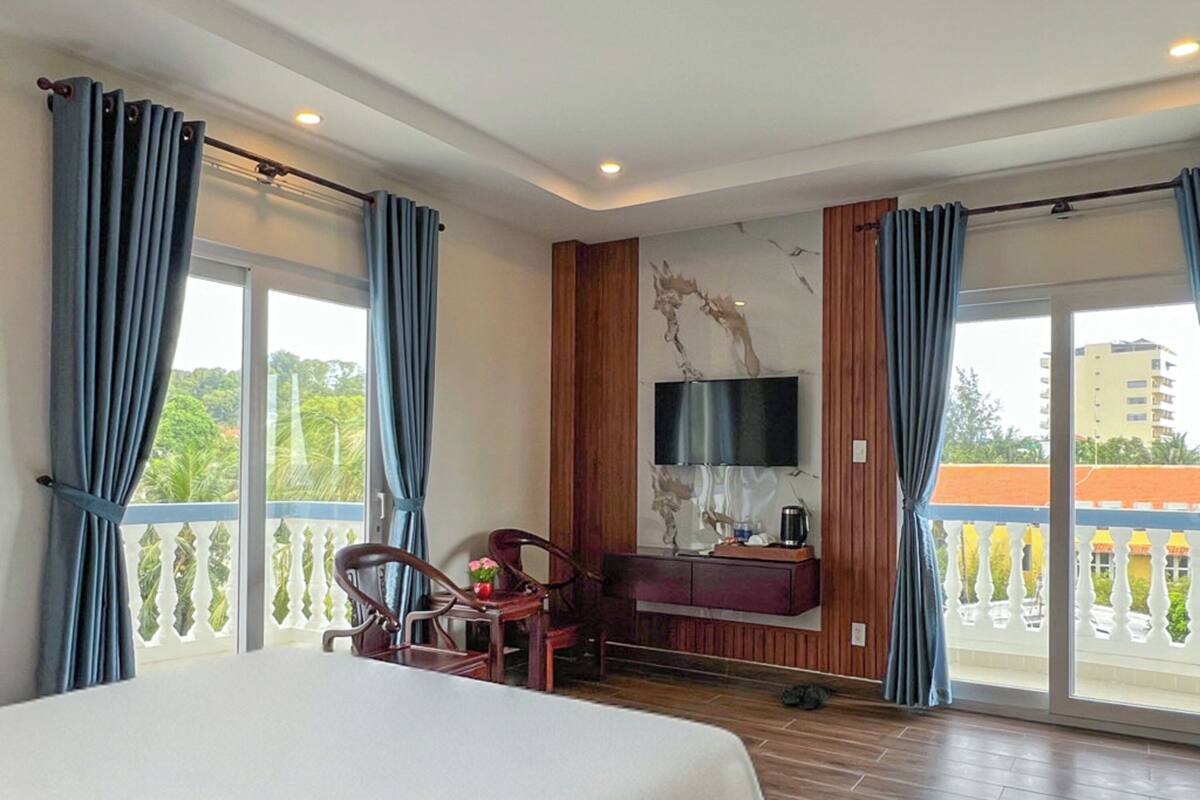 A hotel room with a large bed, a TV mounted on the wall, a wooden desk, and a balcony with curtains.