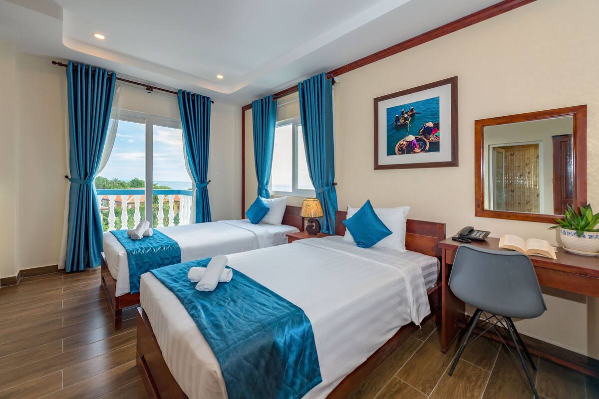 A hotel room with two beds, a desk, a chair, a lamp, a phone, a book, and a window with curtains.
