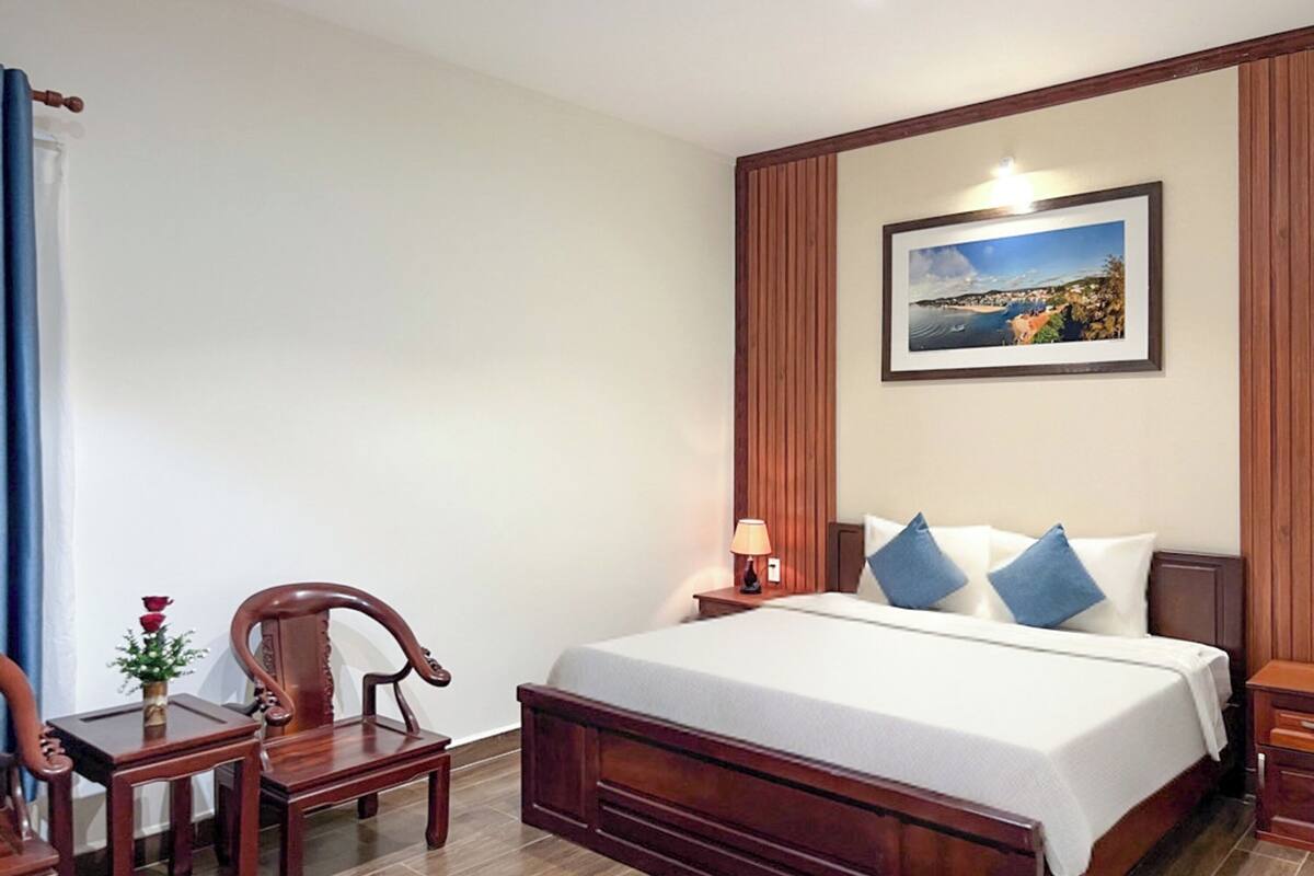 A hotel room with a large bed, a wooden nightstand, a desk with a chair, a lamp, and a framed picture on the wall.