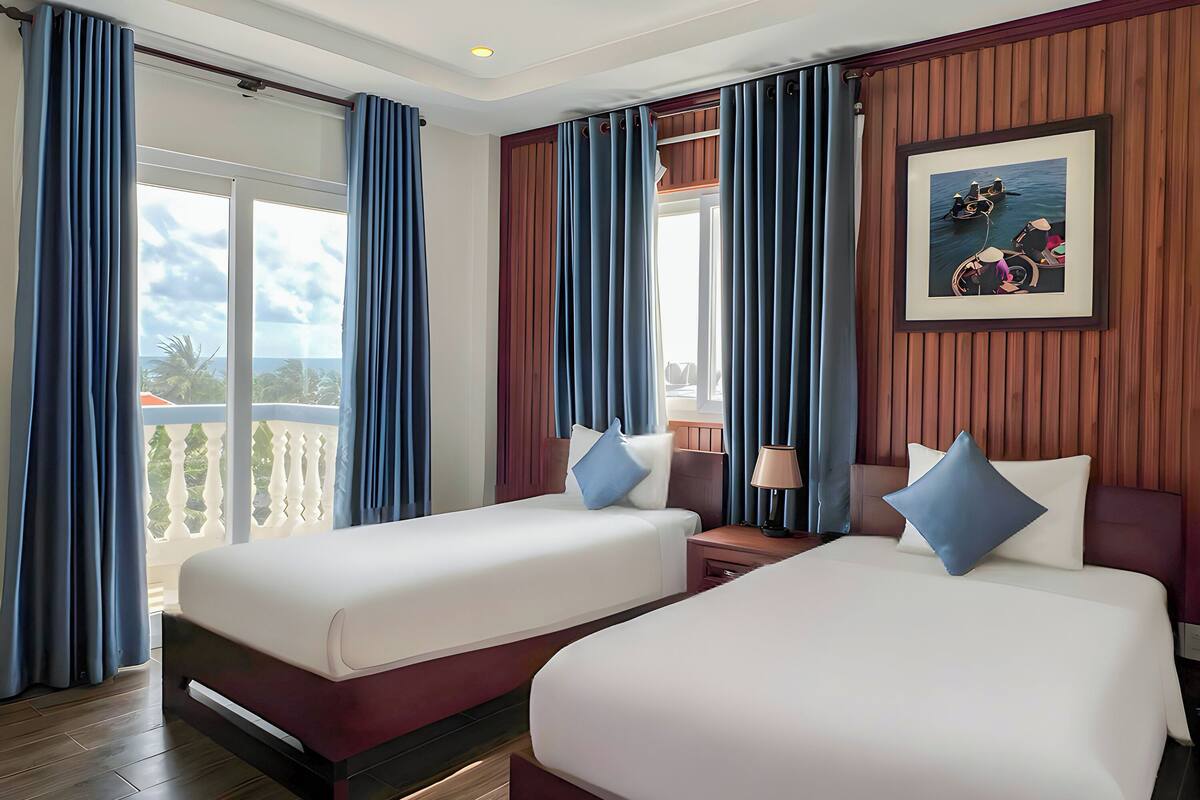 A hotel room with two beds, a large window with curtains, a wooden wall panel, and a framed picture.