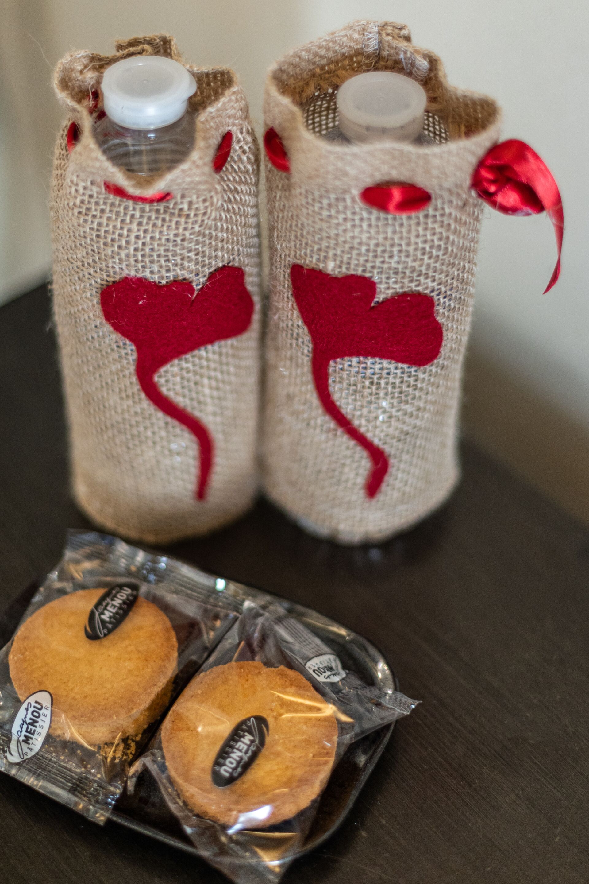Deux bouteilles emballées dans du tissu de jute, ornées de motifs en forme de cœur rouge, et un paquet de biscuits.