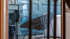 Un escalier avec une rampe en bois mène à une pièce où se trouvent un fauteuil à motifs, une cheminée et un mur bleu orné de moulures décoratives.