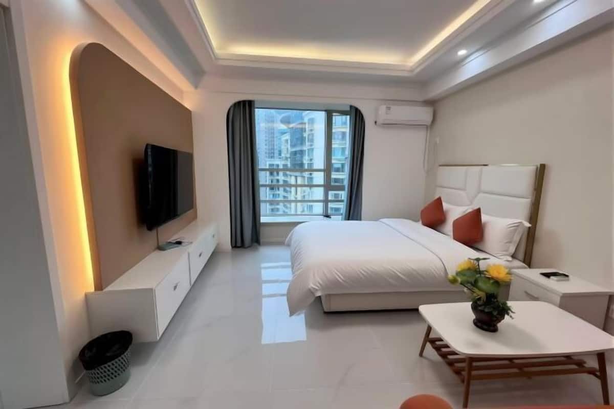 A modern hotel room with a large bed, a TV, a small table with a vase, and a view of the city.
