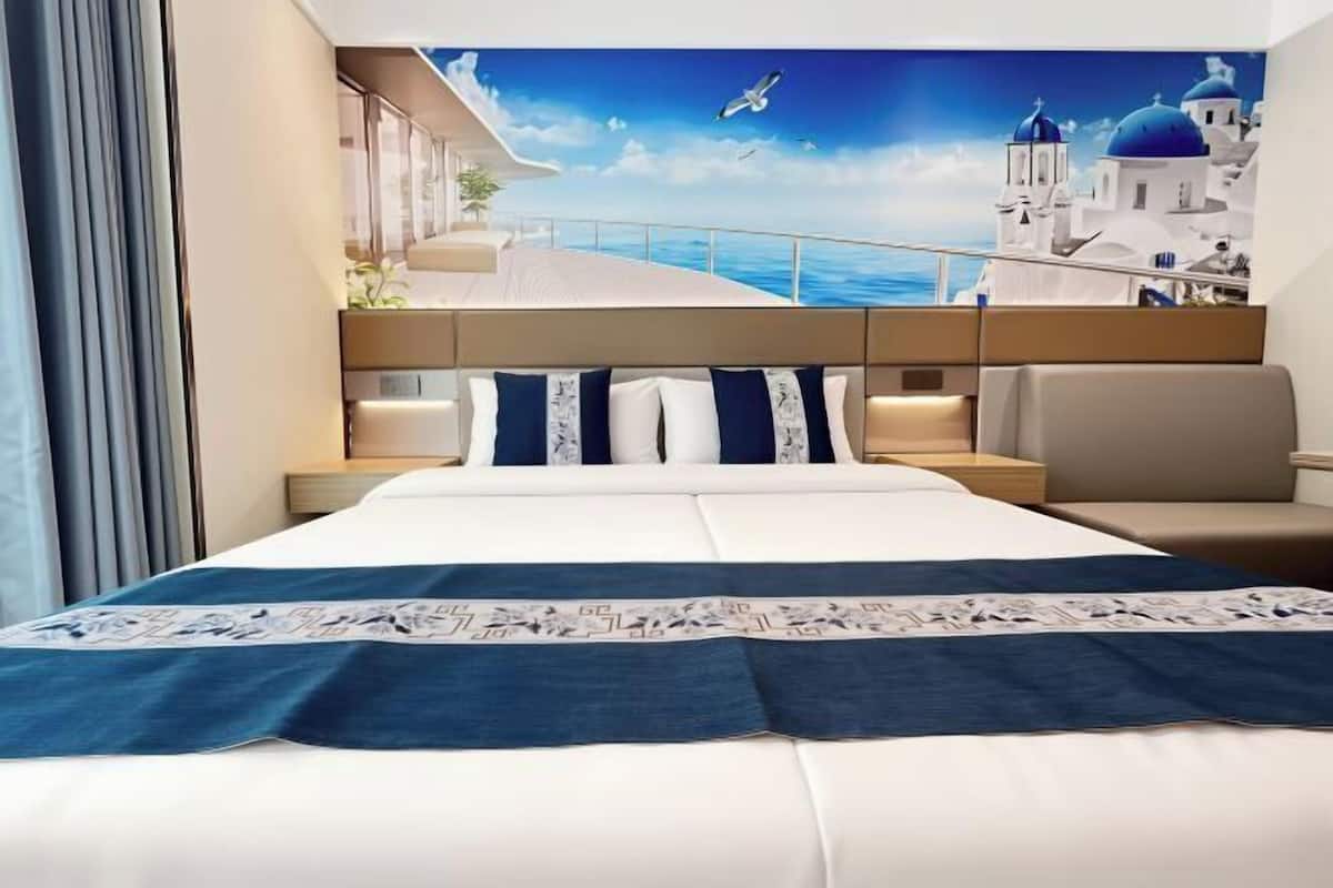 A hotel room with a large bed, a sofa, a nightstand, and a view of a coastal scene with buildings and a blue sky.