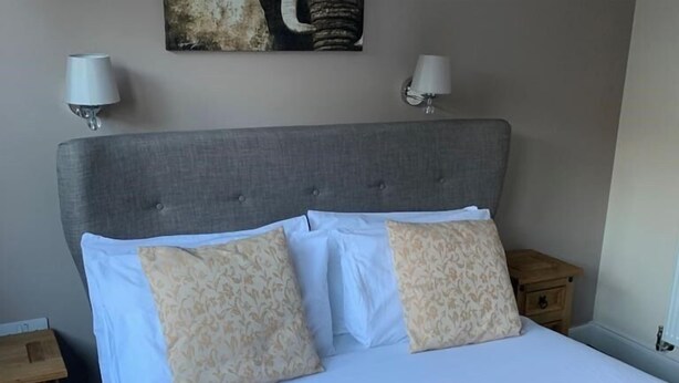 A neatly made bed with a grey upholstered headboard, two matching patterned pillows, and white linens. There are two bedside tables with lamps on either side of the bed.