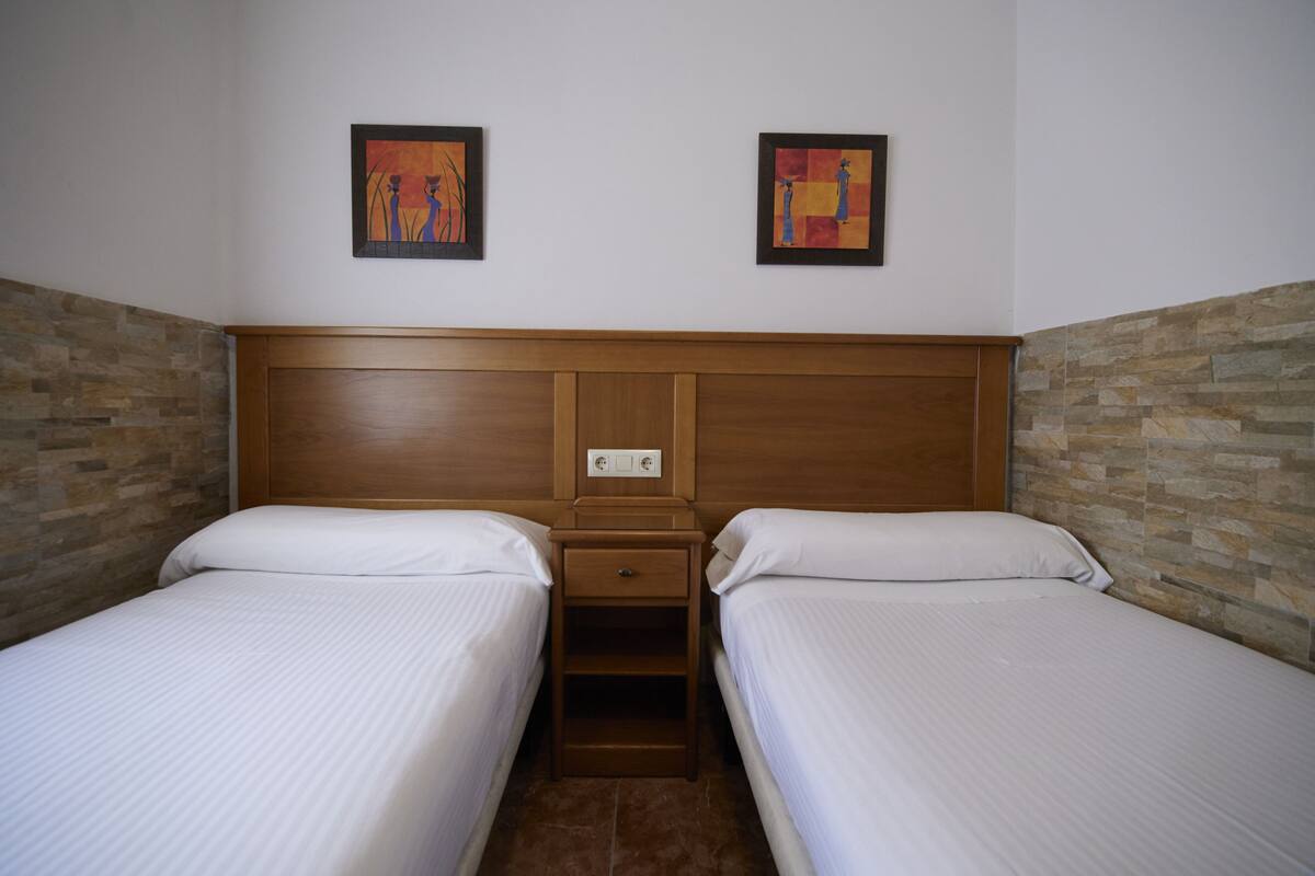 A hotel room with two single beds, a wooden headboard, a nightstand with a drawer, and two framed artworks on the wall.