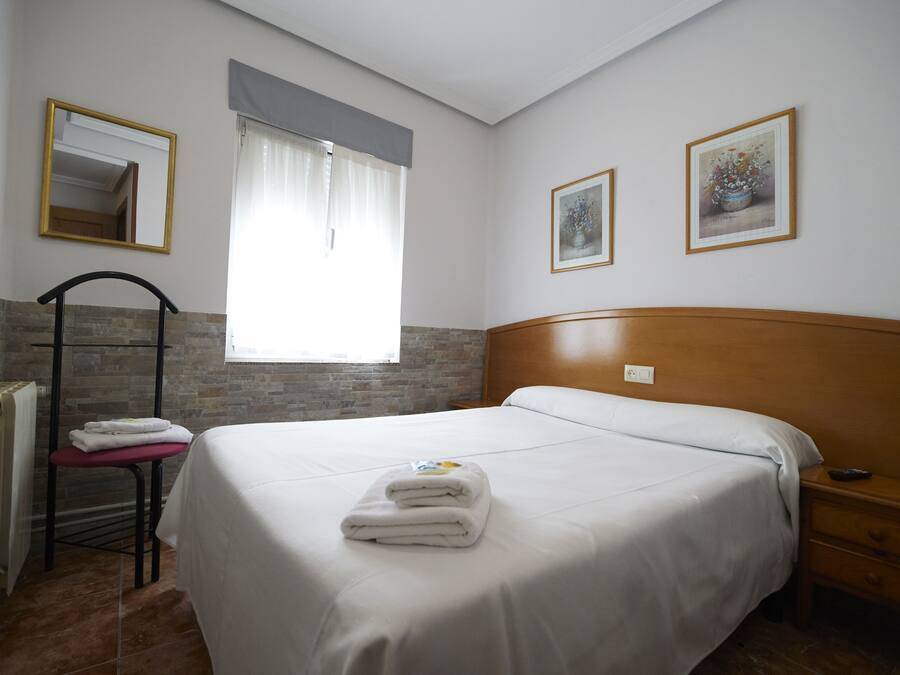 Una cama bien hecha con toallas dobladas, un cabecero de madera, una mesita de noche, un radiador y una ventana con cortinas.