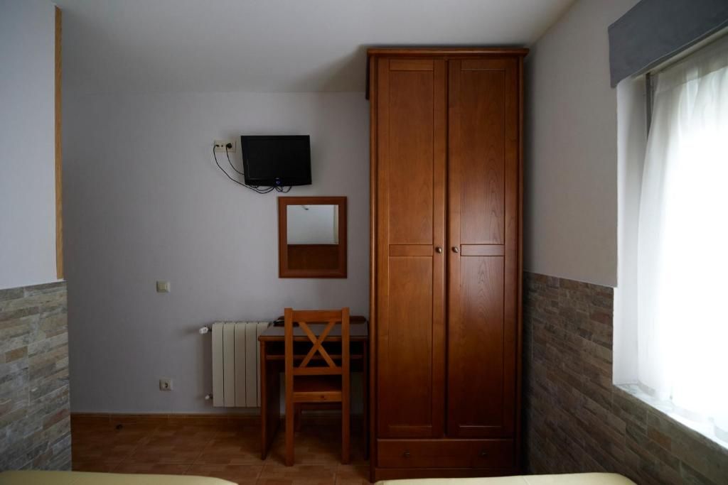 A bedroom with a wooden wardrobe, a desk with a chair, a television mounted on the wall, and a window with curtains.