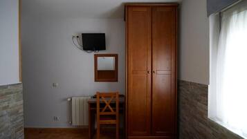 A bedroom with a wooden wardrobe, a desk with a chair, a television mounted on the wall, and a window with curtains.