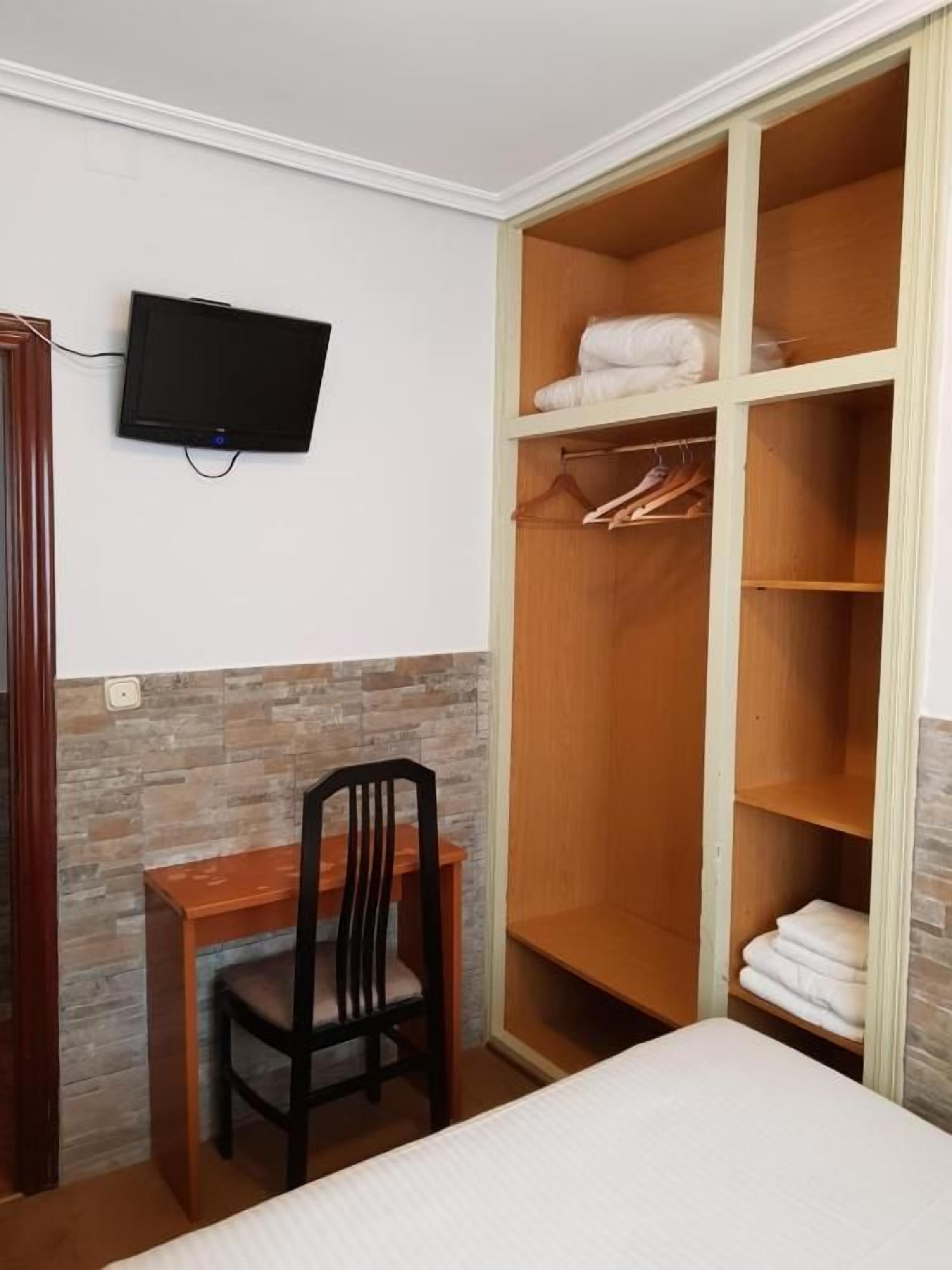 Een slaapkamer met een bed, een bureau met stoel, een aan de muur bevestigde televisie en een houten garderobe met planken en ophanghaakjes.