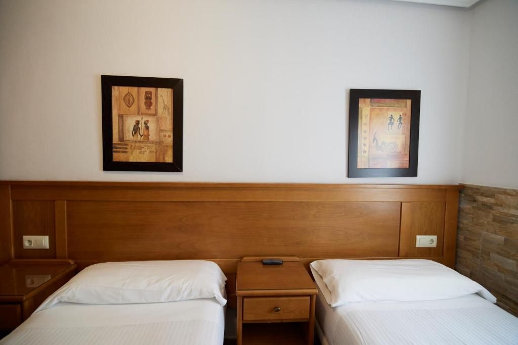 Habitación de hotel con dos camas individuales, cabecera de madera, mesitas de noche y dos cuadros en la pared.