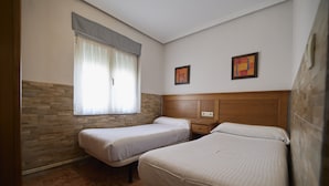 Une chambre avec deux lits simples, une tête de lit en bois, une fenêtre avec des rideaux et deux œuvres d’art encadrées accrochées au mur.