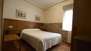 Une chambre à coucher comprenant un lit, une tête de lit en bois, deux tables de chevet, une commode, un petit banc en bois, une fenêtre avec vue et deux tableaux encadrés accrochés au mur.