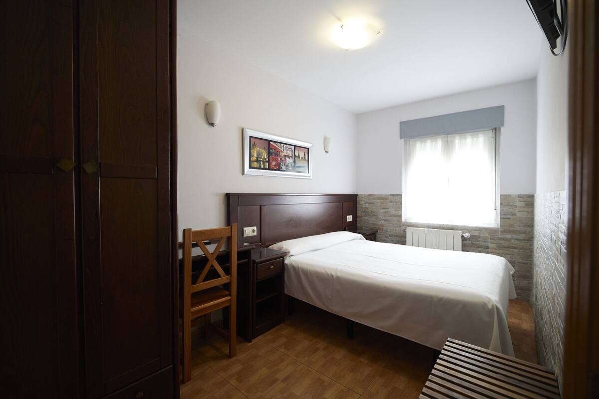 A bedroom with a bed, a desk, a chair, a window with curtains, and a picture on the wall.