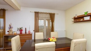 Un comedor con una mesa de vidrio, cuatro sillas, un frutero, un aparador de madera con plantas en macetas y una ventana con cortinas.