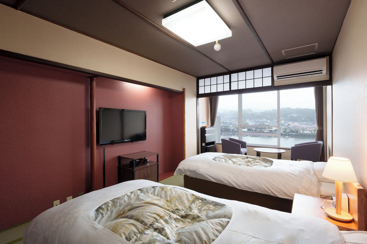 Een hotelkamer met twee bedden, een televisie, een bureau en uitzicht op een stadsgezicht door grote ramen.