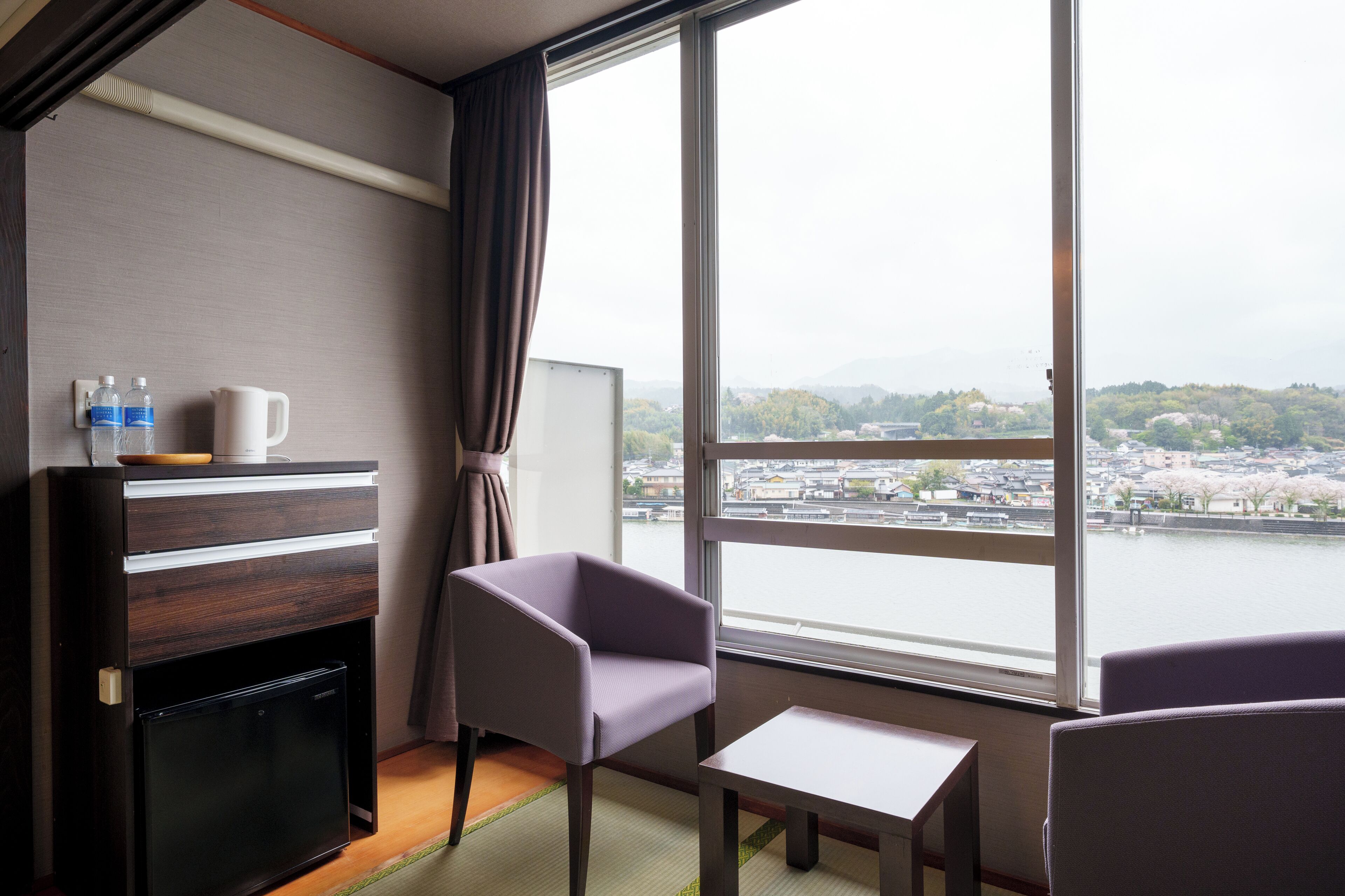 Hotelkamer met uitzicht op een rivier en stadsbeeld, een bureau, twee stoelen, een minibar en een waterdispenser.