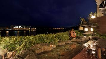 A nighttime view of a waterfront area with a wooden deck, rocks, and a small building with a light.