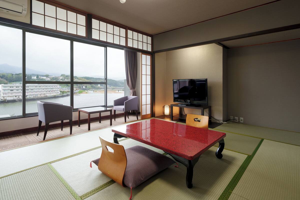 Een kamer met tatami matten, een rode tafel, stoelen, een televisie en uitzicht op een rivier en gebouwen.