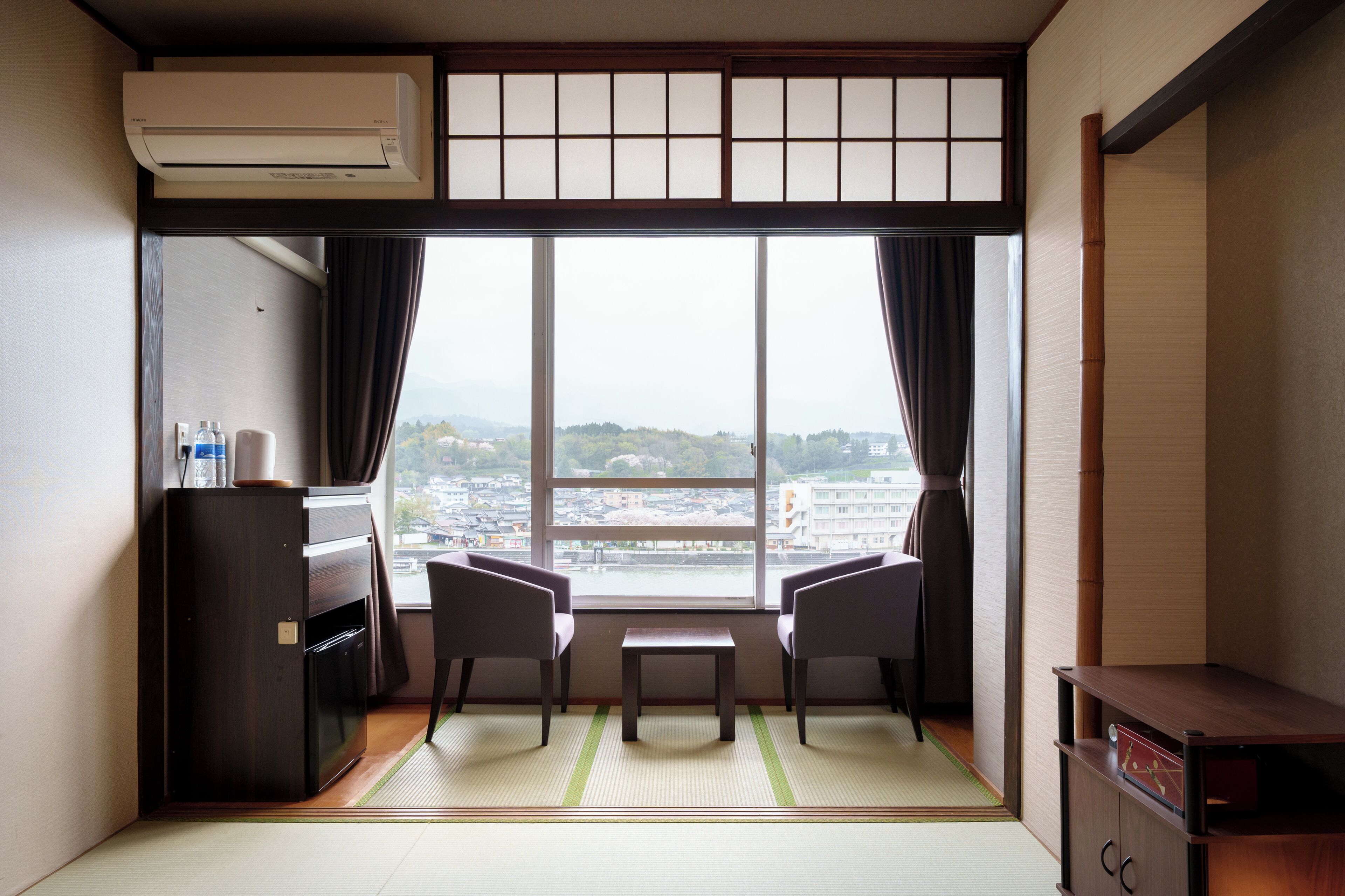A room with tatami flooring, a desk, two chairs, a small table, and a view of a cityscape through large windows.