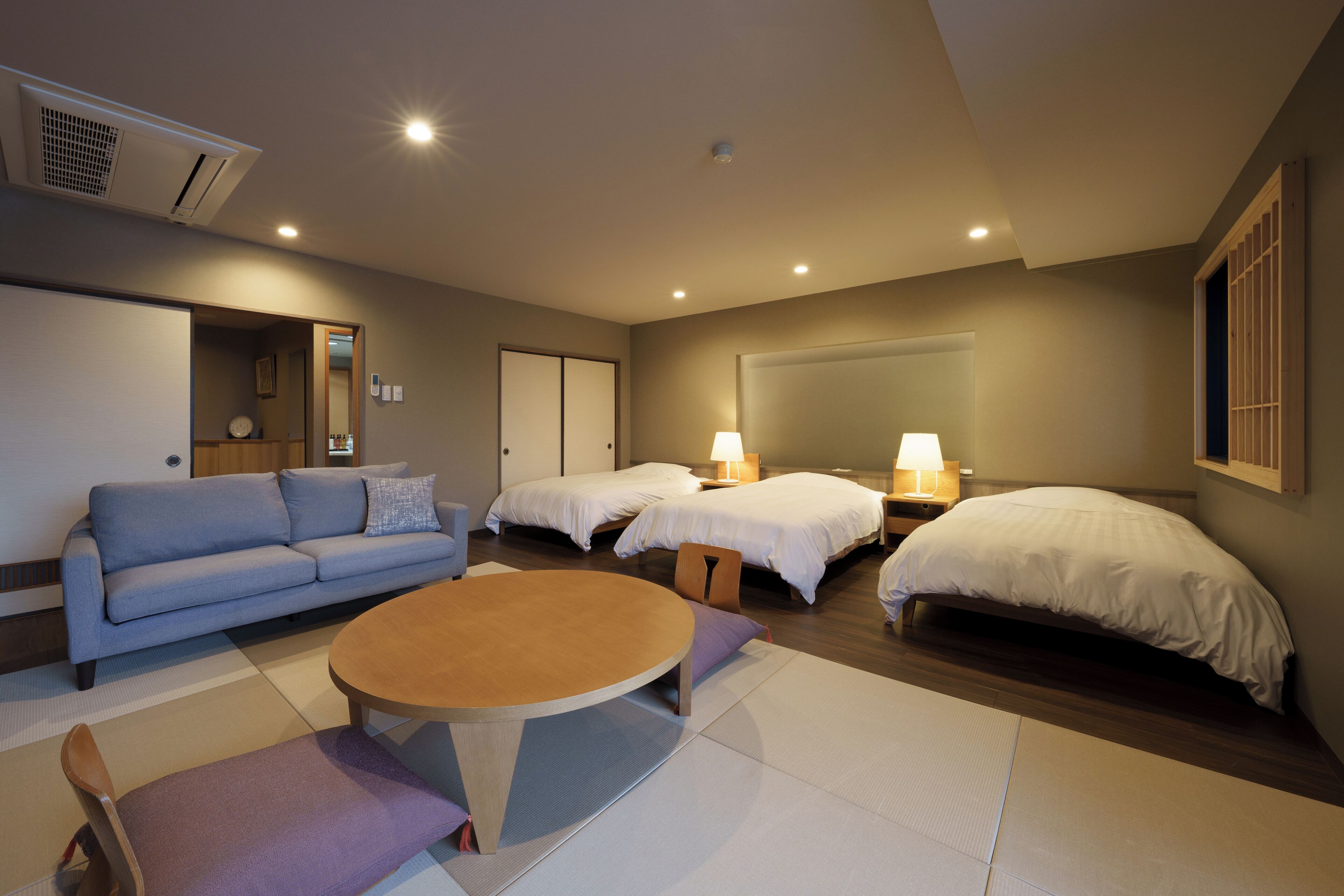 A modern hotel room with a sofa, two single beds, a round table, and a small seating area.