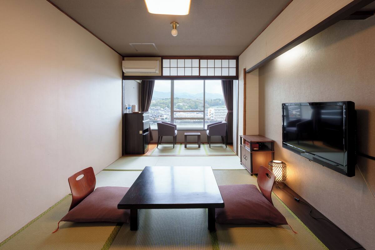 Een traditionele Japanse kamer met tatami vloeren, een lage tafel en kussens. Een televisie is aan de muur bevestigd, en er is uitzicht op een stadsgezicht door grote ramen.