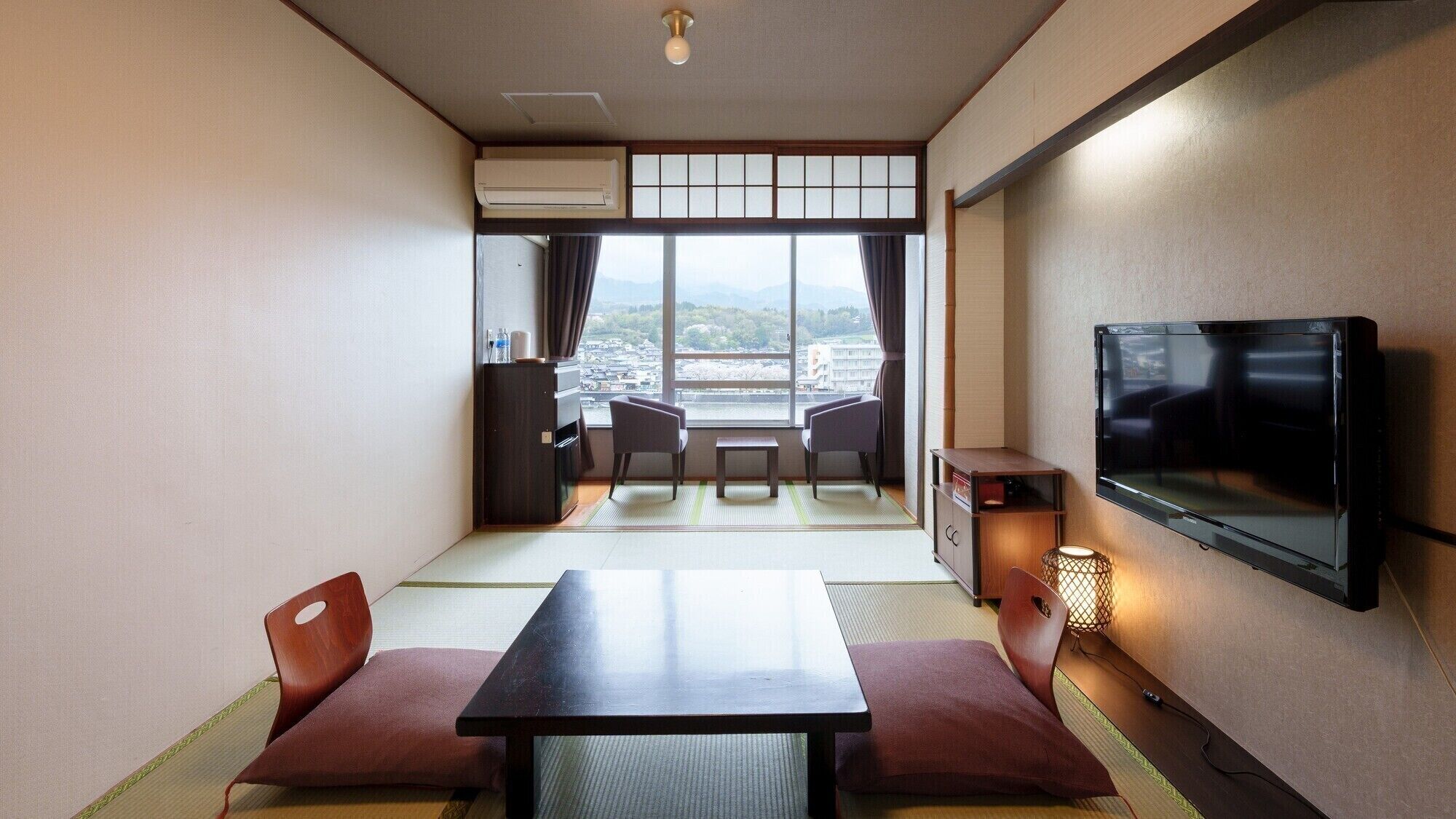 A hotel room with a TV, a dining table, chairs, a sofa, a small table, and a view of the city through large windows.