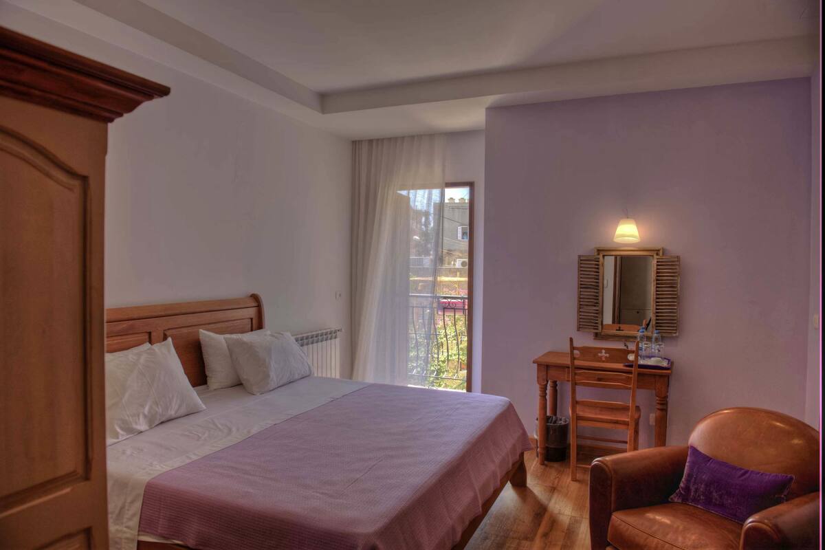 A bedroom with a bed, a desk, a chair, and a window with a view of greenery.