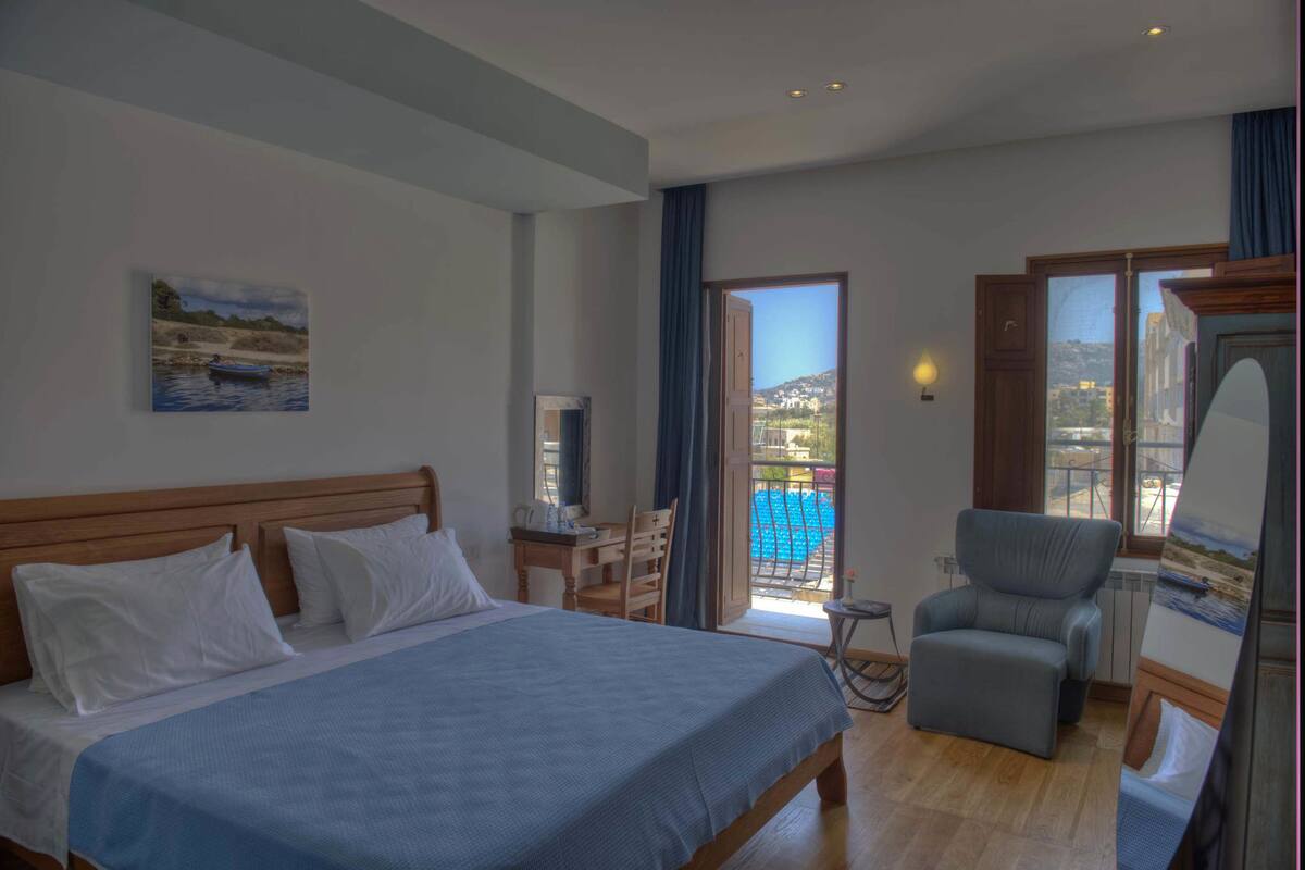 A bedroom with a bed, a desk, a chair, a painting, and a view of a pool and buildings.