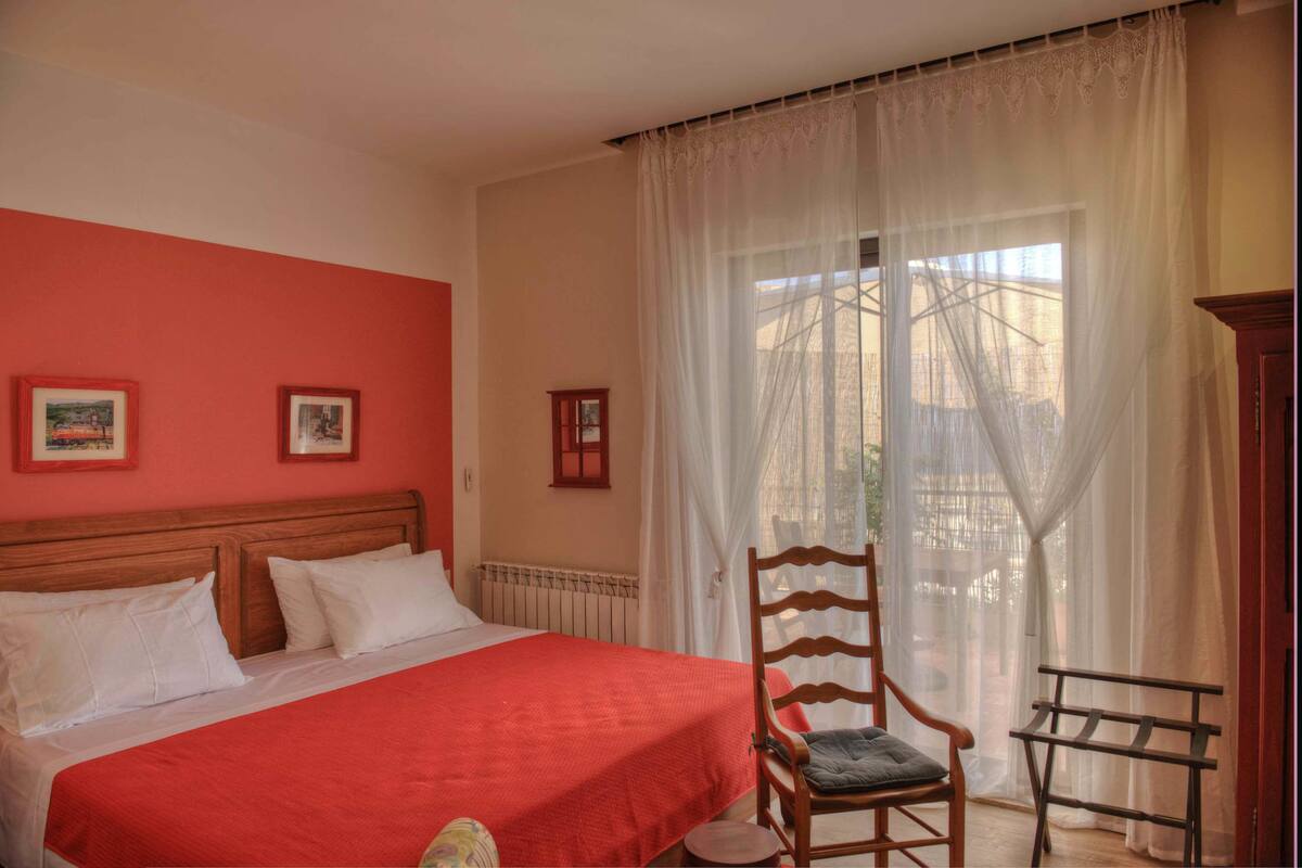 A bedroom with a bed, a chair, a small table, and a window with sheer curtains.