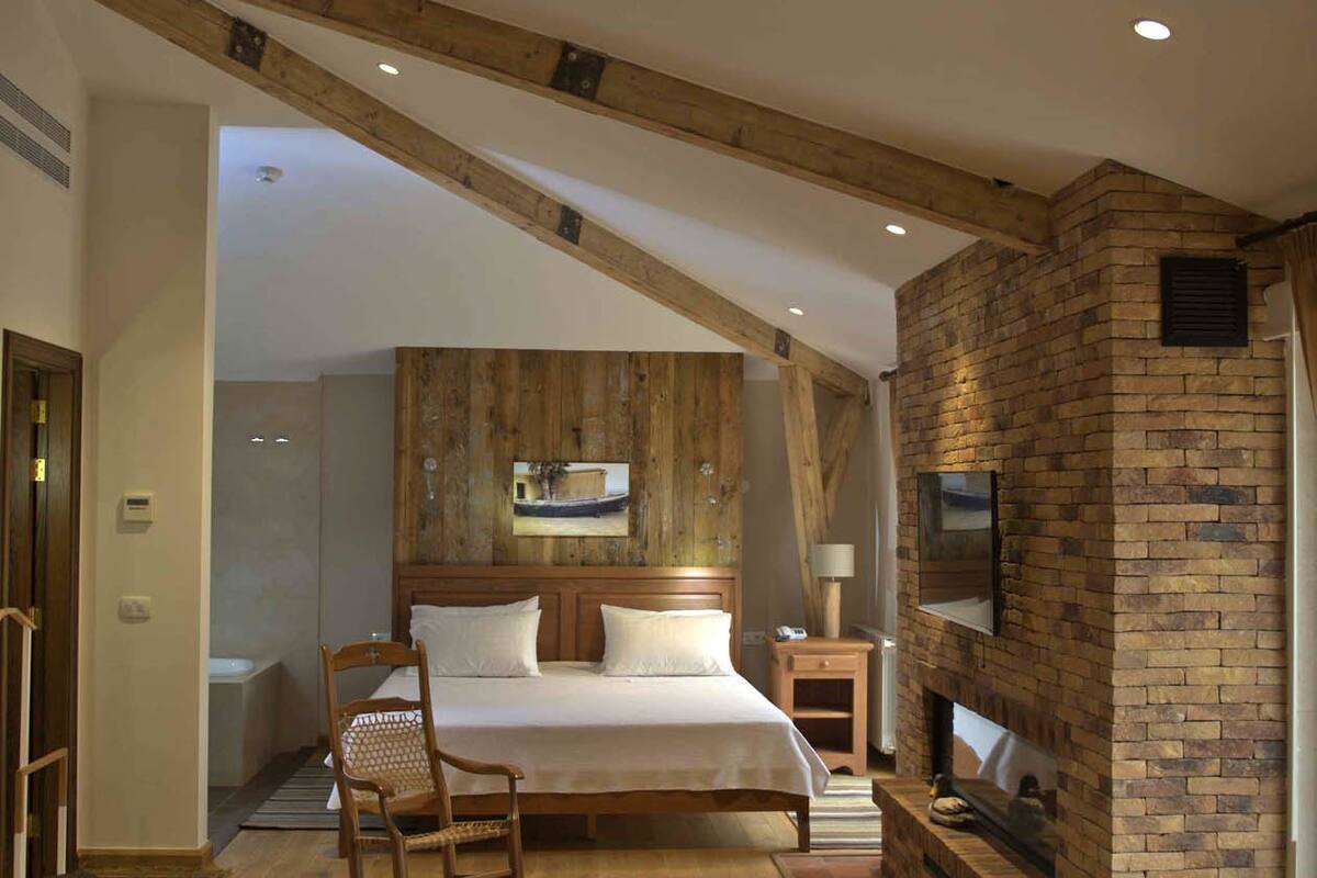 A spacious bedroom with a large bed, wooden beams, a fireplace, and a view of the bathroom.