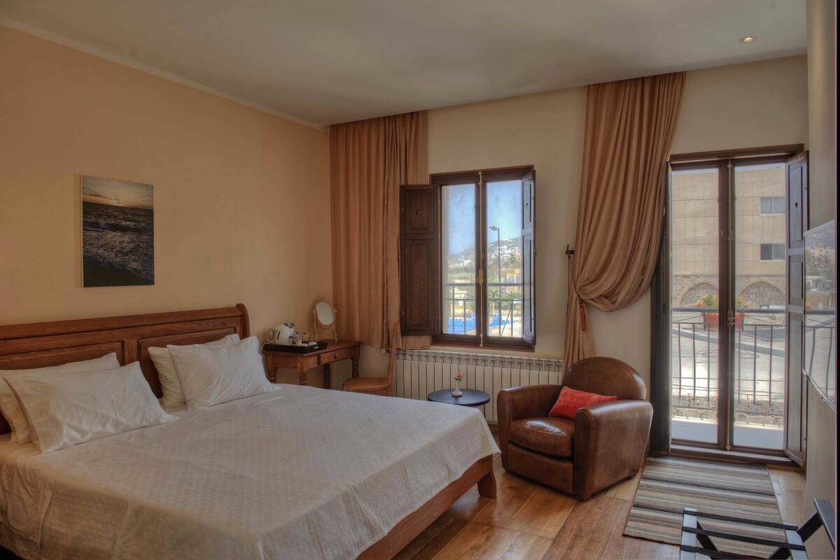 A hotel room with a large bed, a chair, a small table, a window with curtains, and a view of the outside.