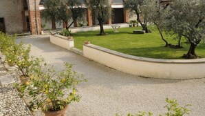 A courtyard with a building featuring arched windows and a tiled roof. There are potted plants and a grassy area. - Villa San Biagio (Colceresa)