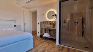 A modern hotel room with a large bed, a glass shower enclosure, a round mirror, and a wooden floor. - Villa San Biagio (Colceresa)