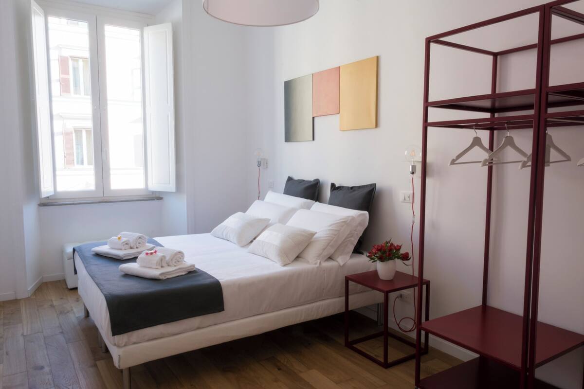 A bedroom with a bed, a bedside table with a vase of flowers, a wardrobe, and a window with blinds.
