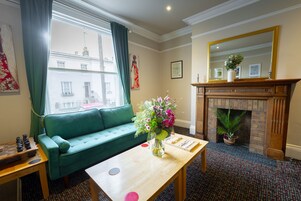 A room with a green sofa, a wooden table with a vase of flowers, a fireplace, and a large mirror.
