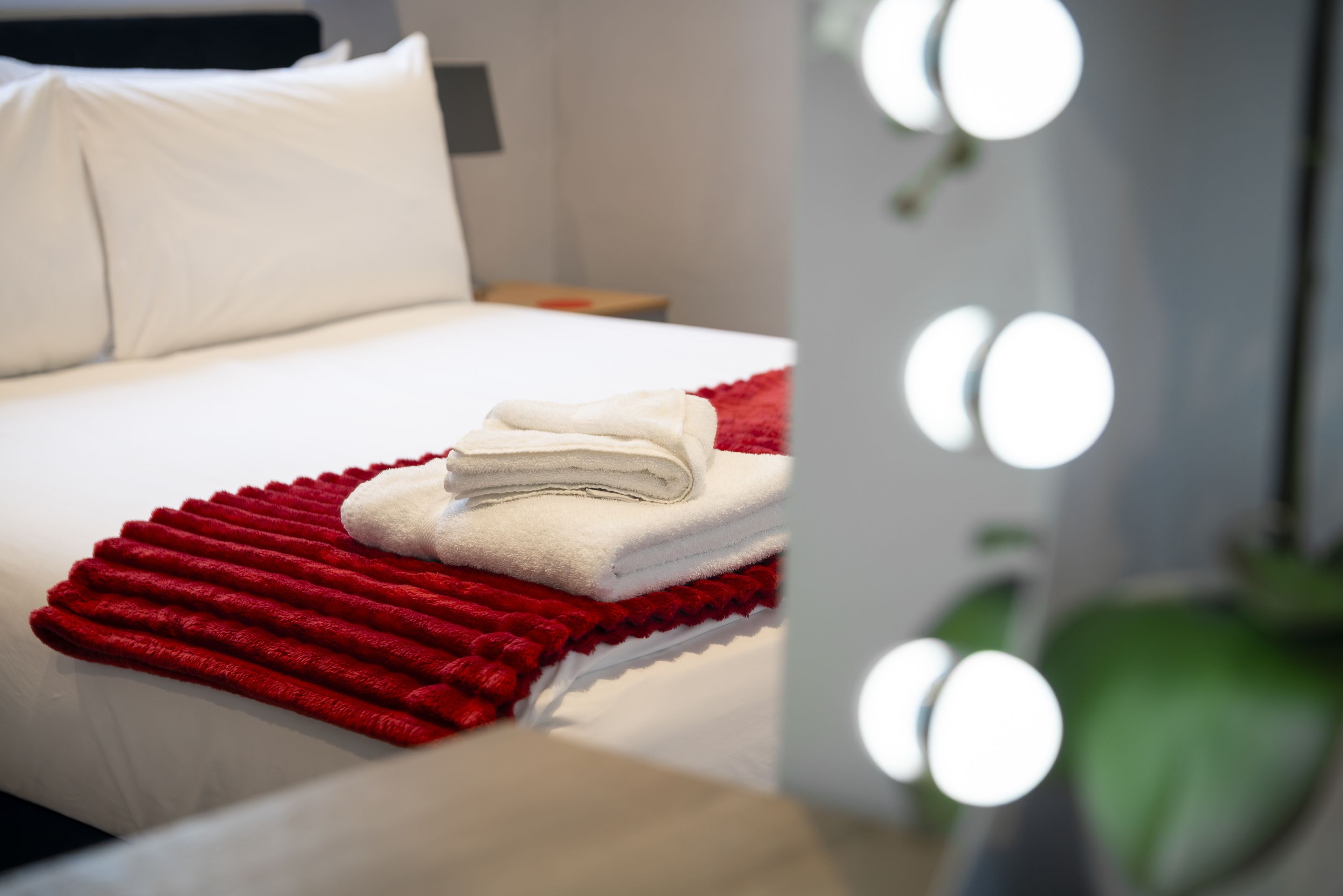 Una cama bien hecha con edredón blanco y una manta roja, un almohadón blanco y una mesita de noche con un objeto rojo encima.