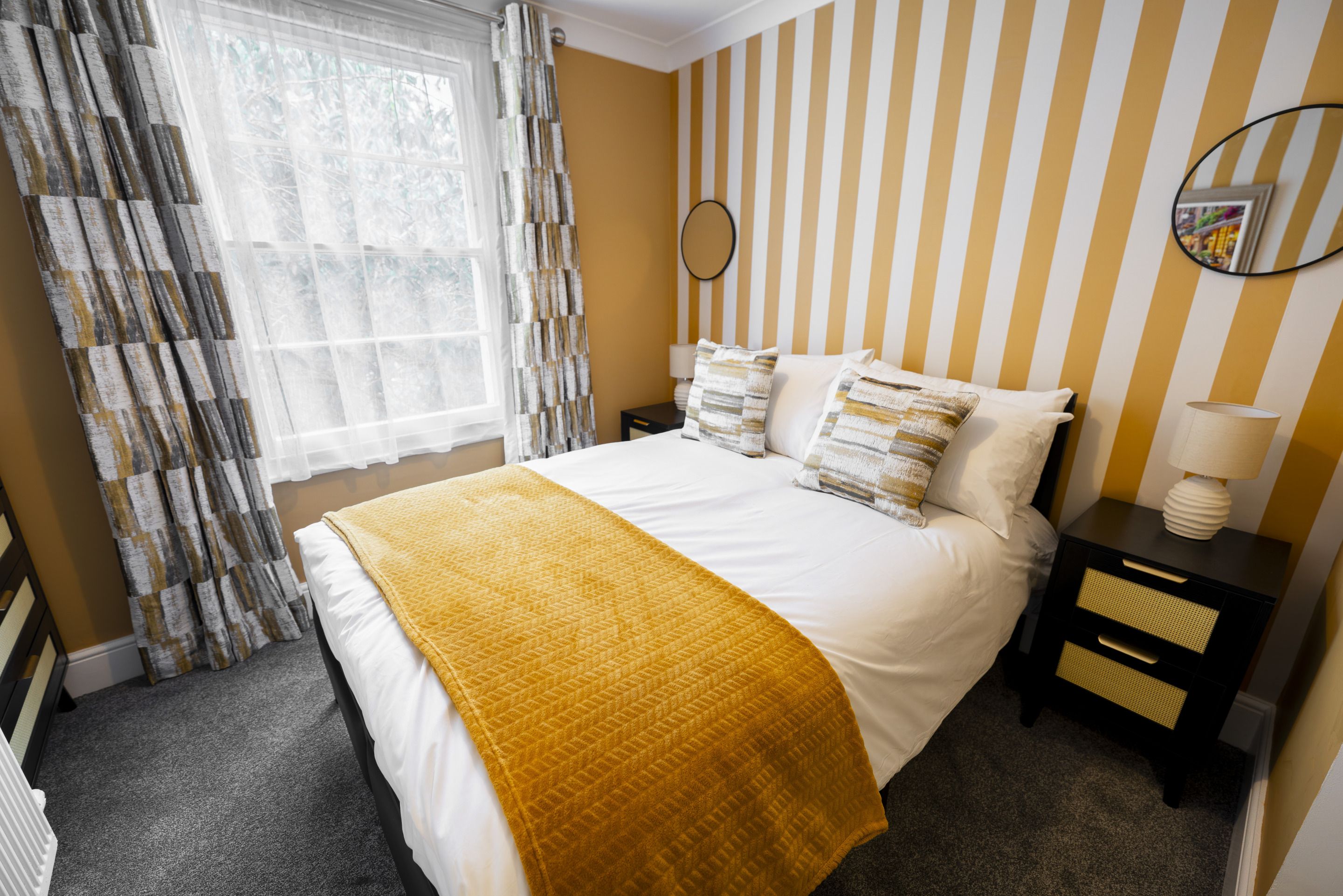 Ein Hotelzimmer mit Bett, Nachttisch, Lampe, Spiegel und einem Fenster mit Vorhängen.