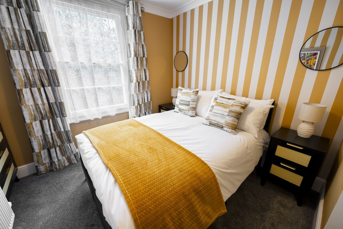 A hotel room with a bed, a bedside table, a lamp, a mirror, and a window with curtains.