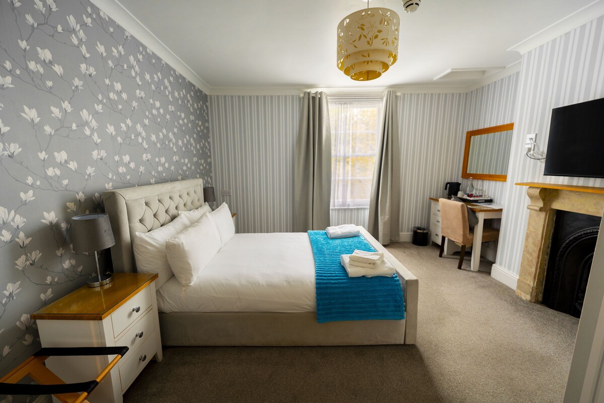 A hotel bedroom with a bed, bedside table, lamp, and a desk with a chair.