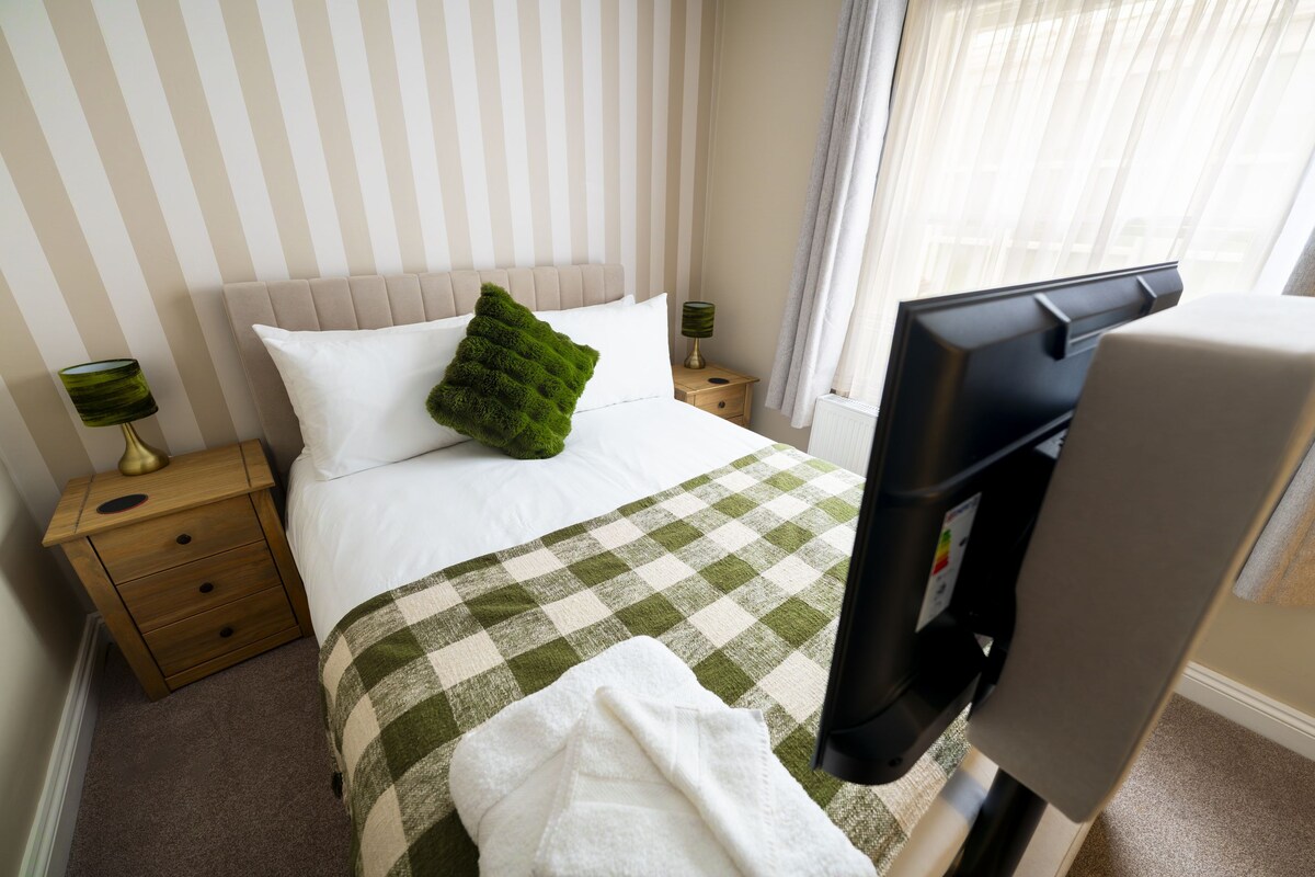 Habitación de hotel con cama, almohada decorativa verde, manta a cuadros, mesita de noche de madera, lámpara y televisión.